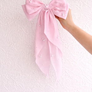May include: A large, light pink bow with long, flowing tails. The bow is adorned with small, scattered pearls and a pearl center. The fabric appears soft and delicate, suitable for hair accessories or decorative purposes.