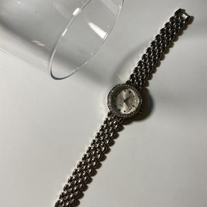 Puede incluir: Un reloj de pulsera plateado con esfera redonda y correa de eslabones. La esfera del reloj está adornada con pequeñas piedras brillantes. Un vaso transparente está en el fondo.
