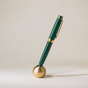 Op de afbeelding: Een donkergroene vulpen met goudkleurige accenten en een clip, rechtop geplaatst in een kleine, bolvormige pennenhouder van geborsteld goudkleurig metaal. De pen heeft een gladde, glanzende afwerking en een goudkleurige ring bij het midden.