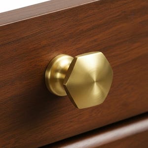 May include: Close-up of a gold-colored, hexagonal cabinet knob attached to a dark brown wooden drawer. The knob has a flat, geometric design and a circular base. The wood grain is visible.