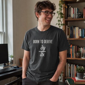 Könnte beinhalten: Ein dunkelgraues Kurzarm-T-Shirt mit dem weißen Text BORN TO DERIVE über einem mathematischen Symbol für die partielle Ableitung dy nach dx. Das T-Shirt hat einen Rundhalsausschnitt und eine Standardpassform aus einfarbigem Stoff.