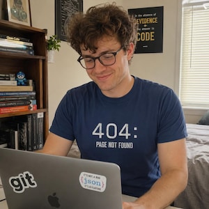 Könnte beinhalten: Ein dunkelblaues T-Shirt mit dem weißen Text 404: PAGE NOT FOUND. Das T-Shirt wird von einer Person mit braunen Haaren und Brille getragen, die auf einen Laptop mit einem git-Logo-Aufkleber und einem Aufkleber IT'S NOT A BUG {json} IT'S A FEATURE schaut.