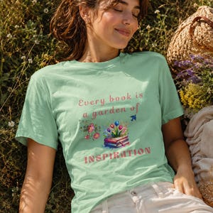 Könnte beinhalten: Ein hellgrünes kurzärmeliges T-Shirt mit dem Schriftzug Every book is a garden of INSPIRATION in rosa und roten Buchstaben, begleitet von einer grafischen Illustration eines Bücherstapels mit bunten Tulpen und floralen Akzenten.