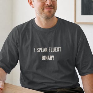 Könnte beinhalten: Ein dunkel anthrazitfarbenes Kurzarm-T-Shirt mit dem Text "I SPEAK FLUENT BINARY", der in einer verwaschenen, cremeweißen serifenlosen Schrift mittig auf der Brust aufgedruckt ist. Das Kleidungsstück hat einen Rundhalsausschnitt.
