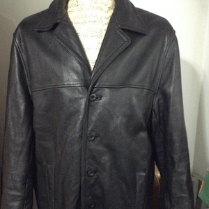 May include: A black leather jacket featuring a classic collar, long sleeves, and a front button closure. The jacket has a smooth texture and a structured fit, with horizontal seam detailing across the chest area and two side pockets.