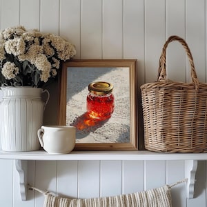 May include: A still-life arrangement on a white shelf. It features a framed painting of a jar of jam, a white ceramic vase with flowers, a small white mug, and a woven basket with a handle. The background is a white paneled wall.