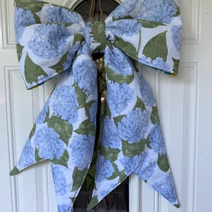 May include: A large fabric bow featuring a repeating pattern of light blue hydrangea flower clusters and green leaves on a white background. The bow has two wide top loops and two long, pointed tails hanging downwards, suspended by a thin twine loop.