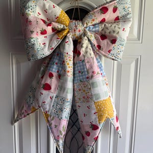 May include: A large decorative bow made from quilted fabric patches featuring various patterns including strawberries, cherries, yellow floral prints, and light blue gingham checks. The bow is suspended by a thin twine loop at the top center.
