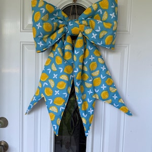 May include: A large fabric bow featuring a repeating pattern of yellow lemons, lemon slices, and small white blossoms on a teal blue background. The bow has two wide top loops and two long, pointed tails hanging down, suspended by a thin brown twine loop.