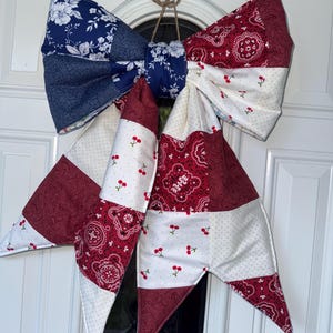 Op de afbeelding: Een grote decoratieve strik van stof, met een patchwork ontwerp in blauw, rood en wit. De strik is versierd met bloemen- en kersenpatronen en wordt opgehangen met een natuurlijke touwlus, geschikt voor deur- of wanddecoratie.