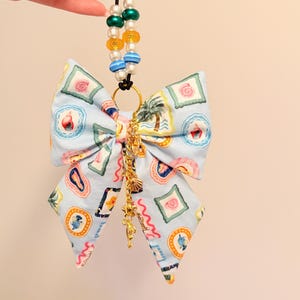 May include: A light blue fabric bow featuring a repeating pattern of tropical motifs including seashells, palm trees, and fish. The bow is attached to a black elastic cord with green, yellow, blue, and white beads, and a gold-toned metal charm chain.