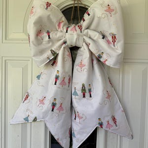May include: A large decorative fabric bow in white, featuring a repeating pattern of nutcracker figures and ballerinas in pink, blue, and red. The bow is suspended by a thin brown twine loop and has two long, pointed tails hanging downwards.