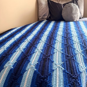 May include: A handmade crochet blanket in shades of blue, white, and navy. The blanket features a textured, striped pattern and is draped over a bed. The color palette creates a calming and cozy aesthetic, perfect for home decor.