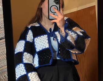 May include: A handmade crochet cardigan in a checkerboard pattern of black, white, and blue. The cropped jacket features a V-neck and long sleeves. The trim and edges are a vibrant blue, adding a pop of color to the design. The cardigan is a unique, cozy garment.