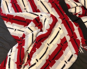 May include: A crocheted blanket featuring a repeating geometric pattern in red, white, light gray, and black yarn. The design consists of diagonal stripes with textured, raised stitches and a fringe border made of white and red yarn strands.