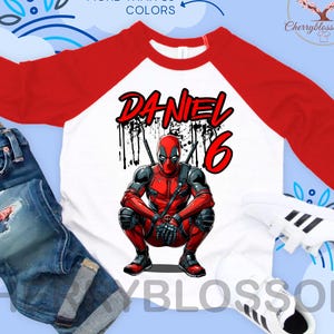 May include: A white and red raglan shirt featuring a Deadpool graphic with the name "DANIEL" and the number "6". The shirt is paired with blue jeans and white sneakers. The text "MORE THAN 10 COLORS" is visible.
