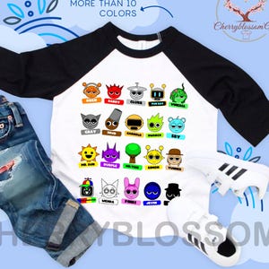 May include: White and black raglan sleeve shirt with colorful cartoon character graphics. The shirt features the text "MORE THAN 10 COLORS" and is paired with blue jeans and white sneakers.