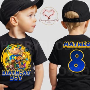 May include: Black t-shirt with a cartoon graphic and the text "BIRTHDAY BOY" in blue and yellow. The back of the shirt has the name "MATHEO" and the number "8" in blue and yellow. The child is wearing a black baseball cap.