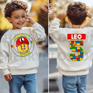 May include: A cream-colored sweatshirt with a Lego-themed birthday design. The front features a Lego character with the text "BIRTHDAY BUILDER". The back displays the name "LEO" and the number "5" in a Lego brick pattern. The sweatshirt is paired with blue jeans.