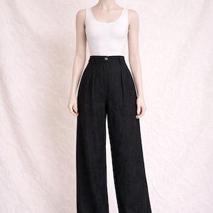 Peut inclure: Un mannequin porte un débardeur blanc et un pantalon noir à taille haute et à jambes larges. Le pantalon a une fermeture boutonnée et des plis à la taille. Le fond est un tissu rose clair.