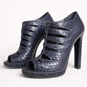 Könnte beinhalten: Ein Paar dunkel marineblaue High-Heels mit texturiertem Schlangenledermuster. Diese Peeptoe-Stiefeletten verfügen über eine Reihe horizontaler Ausschnitte an den Seiten und vorne, eine Plateausohle sowie einen hohen, dicken Blockabsatz.