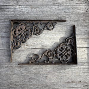 May include: Two ornate, cast iron shelf brackets with a dark brown, aged finish. Each bracket features a floral design and scrollwork, perfect for adding a vintage touch to any space. The brackets are approximately 8 inches long.
