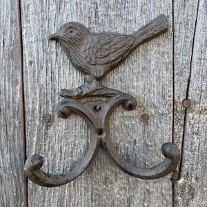 May include: A cast iron wall hook featuring a detailed bird perched above two curved hooks. The bird and hooks have a rustic, dark brown finish. The hook is mounted on a weathered wooden surface.
