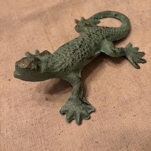 May include: A cast metal lizard figurine in a distressed teal finish with bronze accents. The lizard is posed with its legs spread and tail curled. The figurine is approximately 6 inches long.