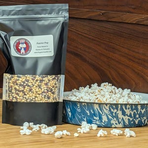 May include: A black bag of "Patriot Pop" popcorn with a clear window showing kernels, next to a blue and white enamel bowl filled with popped popcorn. Kernels and popped popcorn are scattered on a wooden surface.