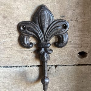 May include: A cast iron fleur-de-lis wall hook with a dark, textured finish. The hook features the classic three-petal design, with a decorative stem and a rounded hook end. It is mounted on a wooden surface.