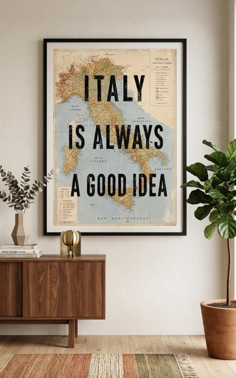 May include: A framed map of Italy with the text "ITALY IS ALWAYS A GOOD IDEA" in bold black letters. The map is in shades of tan and blue, and the frame is black. The artwork is displayed on a white wall.