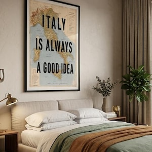 May include: A framed poster with the text "ITALY IS ALWAYS A GOOD IDEA" over a vintage map of Italy. The poster hangs above a bed with white pillows, a green duvet, and an orange throw blanket. A lamp and side table are visible.