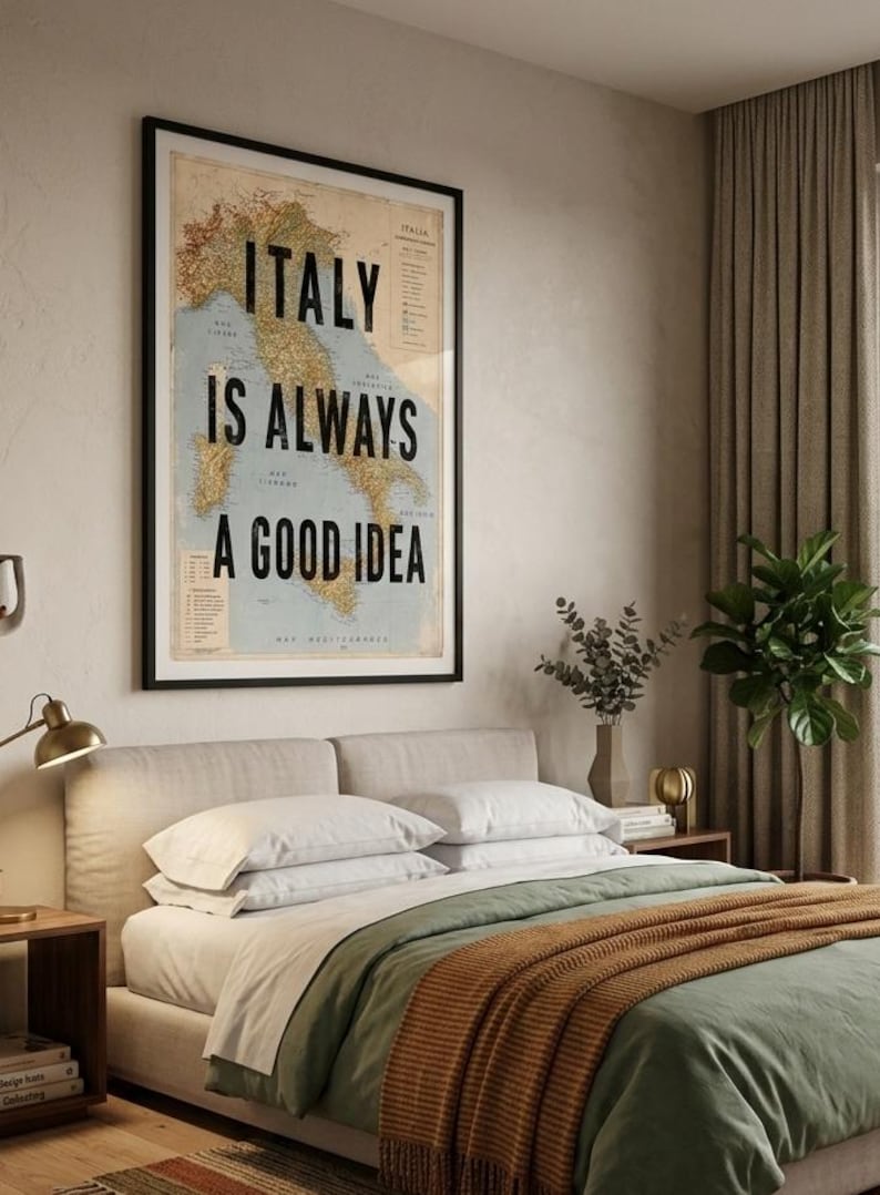 May include: A framed poster with the text "ITALY IS ALWAYS A GOOD IDEA" over a vintage map of Italy. The poster hangs above a bed with white pillows, a green duvet, and an orange throw blanket. A lamp and side table are visible.