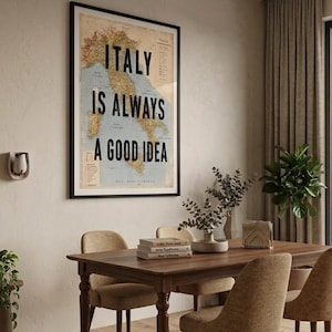 May include: Framed wall art with the text "ITALY IS ALWAYS A GOOD IDEA" over a vintage map of Italy. The artwork hangs above a wooden dining table with chairs, books, and decorative items.