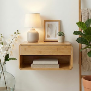May include: A light wood floating nightstand with a drawer and open shelf. A beige lamp, framed artwork, and a small potted succulent sit on top. Two books are on the shelf below. A vase of white flowers is in the foreground.