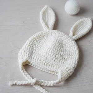 Puede incluir: Un gorro de bebé blanco tejido a ganchillo con orejas de conejo y un pompón a juego. El gorro presenta una corona redondeada, solapas para las orejas y lazos trenzados. Las orejas de conejo están erguidas y añaden un toque lúdico. El pompón es una bolita pequeña y esponjosa.