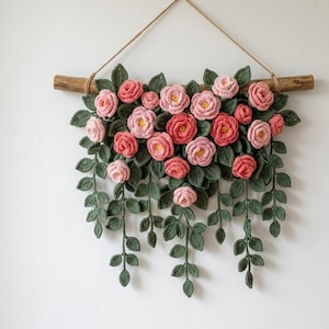 Puede incluir: Un colgante de pared hecho a mano con rosas de ganchillo en tonos rosa y rojo, con centros amarillos. Las flores están unidas a hojas y enredaderas verdes de ganchillo, suspendidas de un pasador de madera con un colgador de hilo natural. Decoración floral.
