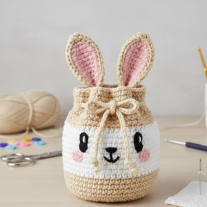 May include: A handmade crochet bunny bag in beige, white, and pink. The bag features a drawstring closure, bunny ears, and a cute face with black eyes, a black nose, and pink cheeks. The bag is likely used for storing small items.