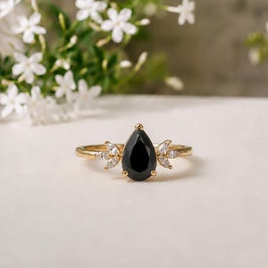 May include: A gold ring featuring a central pear-shaped black gemstone held by prongs. The band is accented on each side by a cluster of three small, clear marquise-cut stones arranged in a fan-like pattern, creating a symmetrical design.