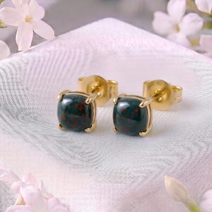 May include: A pair of stud earrings featuring dark green, rounded stones with small red speckles, set in polished gold-tone metal settings. The earrings have standard post backs and are displayed on a textured white fabric surface.