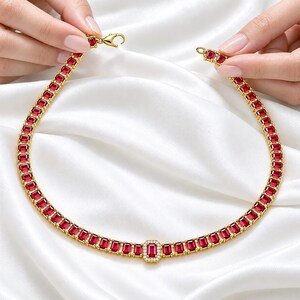 May include: A gold necklace featuring a continuous row of rectangular red gemstones. The centre of the necklace showcases a larger rectangular red gemstone surrounded by a halo of small white stones. It includes a gold lobster claw clasp.