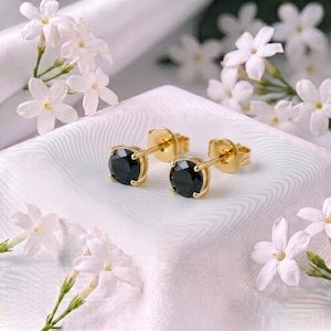 May include: A pair of gold stud earrings featuring round, faceted black stones set in a four-prong gold setting. The earrings are displayed on a white fabric surface surrounded by small, white five-petaled flowers with green stems.