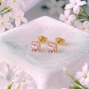 May include: A pair of stud earrings featuring square-cut, pale peach-coloured gemstones set in gold-toned metal. The earrings are displayed on a textured white fabric surface, surrounded by several small, white, five-petalled jasmine flowers.