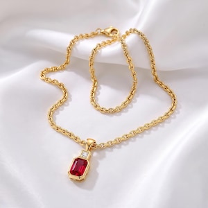 Puede incluir: Un collar de cadena dorada con un colgante de piedra roja de corte esmeralda rectangular, acentuado con una pequeña piedra clara de corte redondo en la parte superior. El colgante cuelga de una cadena de eslabones clásica, creando una pieza de joyería refinada.