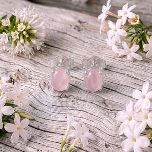 Peut inclure: Une paire de boucles d'oreilles à clous avec des pierres cabochons ovales, lisses et rose pâle, serties dans un métal argenté poli. Les boucles d'oreilles sont placées au centre, entourées de grappes de petites fleurs blanches en étoile.