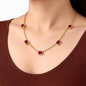 Peut inclure: Un collier en chaîne dorée orné de cinq pierres précieuses rectangulaires rouges espacées, chacune entourée d'un halo de petites pierres blanches. Le collier repose sur un haut côtelé marron foncé, présentant un design à maillons classiques.