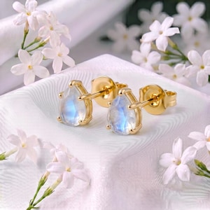 May include: A pair of gold stud earrings featuring pear-shaped moonstones with a blue iridescent glow. The earrings have a prong setting and include gold butterfly backings. The stones are translucent with a smooth, polished surface finish.