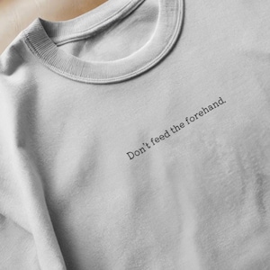 Peut inclure: Un sweat-shirt gris clair avec un col rond. Le texte "Don't feed the forehand" est imprimé en noir. Le sweat-shirt est fabriqué dans une matière douce et confortable.