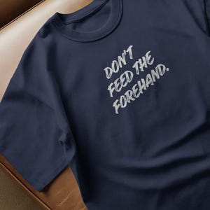 Peut inclure: T-shirt bleu marine avec l'inscription blanche "DON'T FEED THE FOREHAND" dans un style de coup de pinceau. Le t-shirt a un col rond et est fait d'une matière douce.