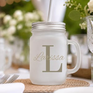 May include: A frosted glass mason jar mug with a silver metal lid and a white straw. The front of the mug features a large, dark beige capital letter L with the name Larissa written in a cursive script font overlaid across the letter.